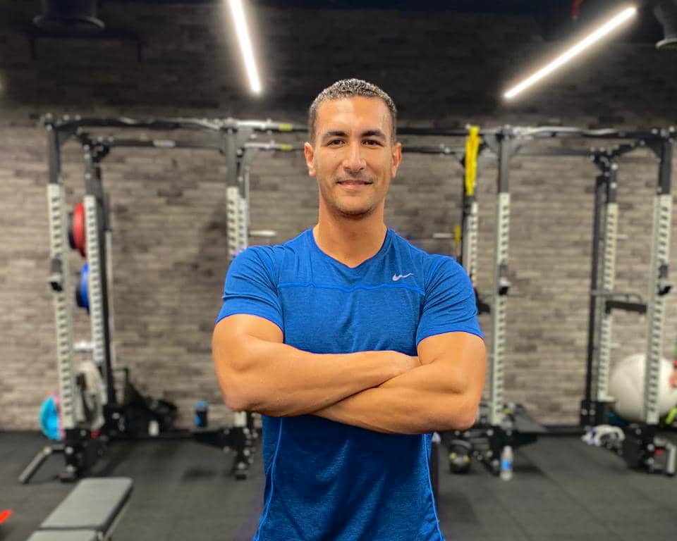 a man wearing blue t-shirt standing in the gym with his hands crossed over chest and he is smiling