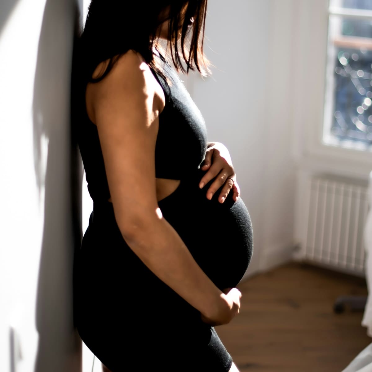 Side profile of a pregnant woman in a black dress leaning against a wall in a sunlit room, illustrating the journey to Stay Active During Pregnancy.