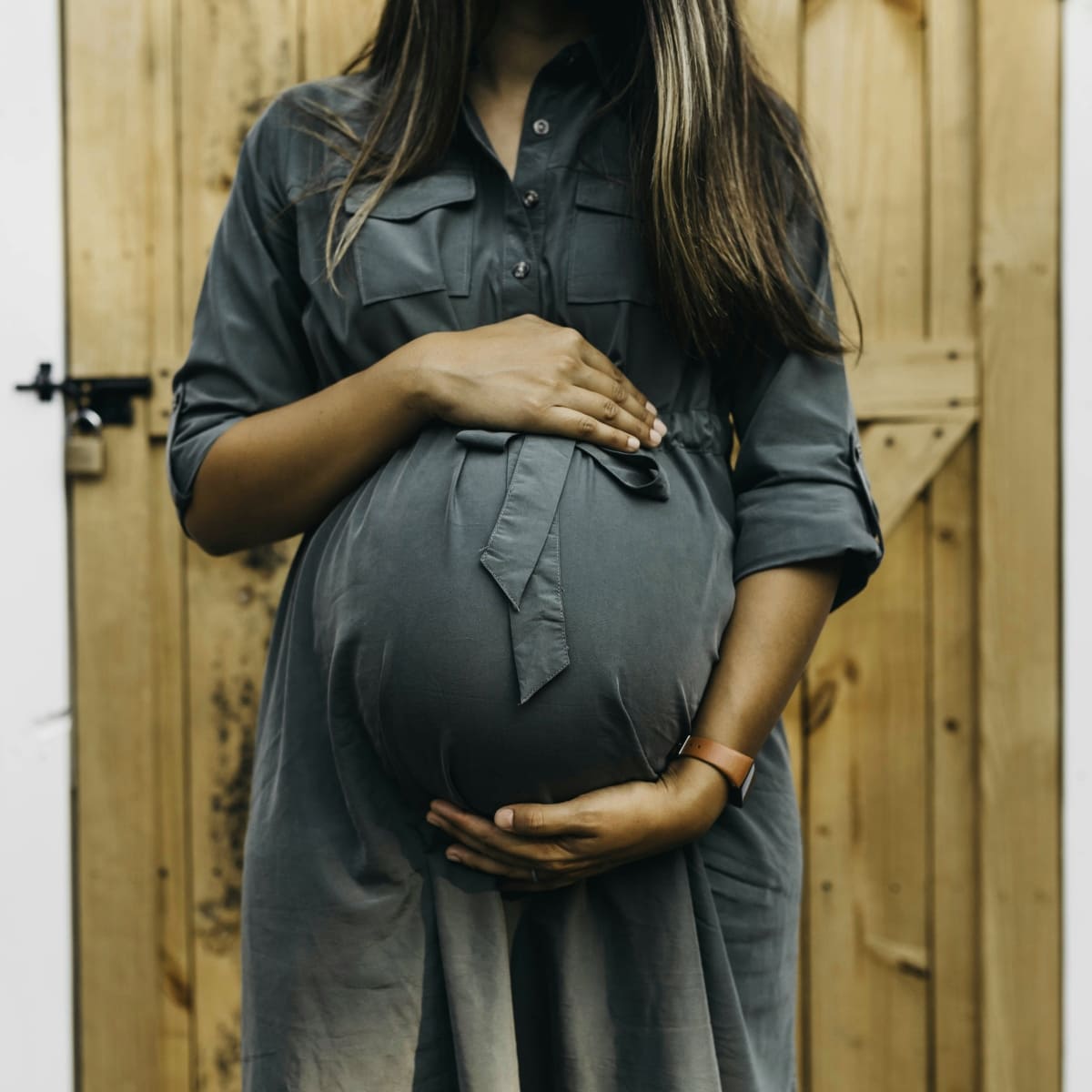 Close-up of a pregnant woman standing in front of a wooden door, cradling her baby bump in a grey dress to highlight ways to Stay Active During Pregnancy.