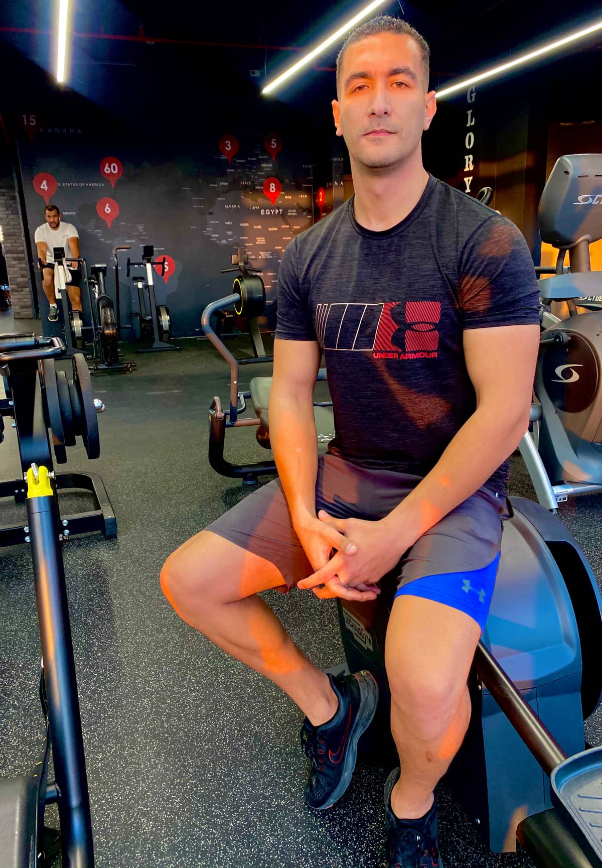 Coach fathi, an Arabic speaking online personal trainer, sitting on a stationary bike in a gym wearing an Under Armour shirt